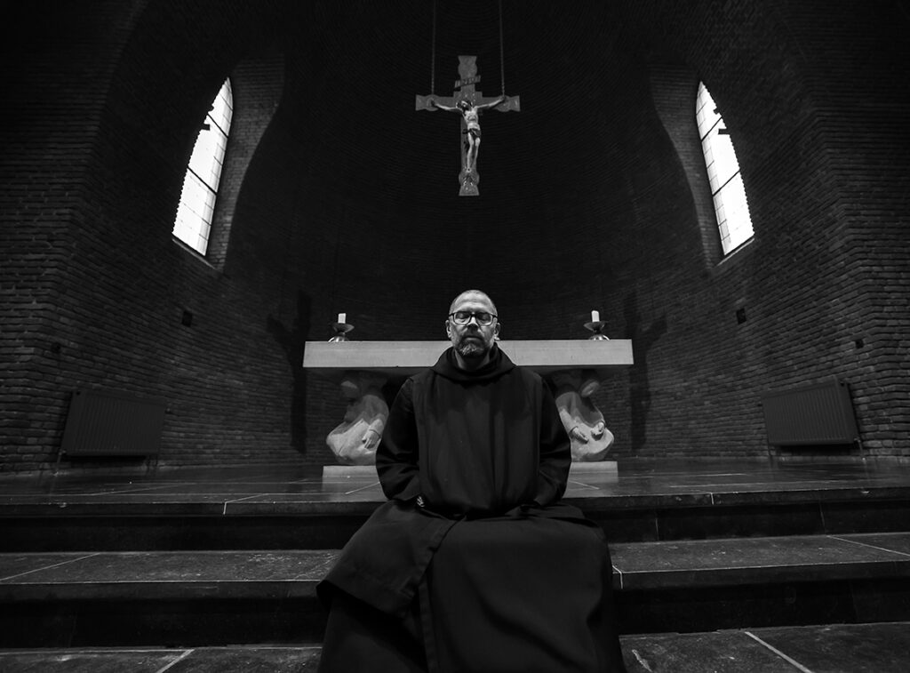 Foto cursist: zwart wit foto van een priester voor een altaar met een groot kruis op de achtergrond.